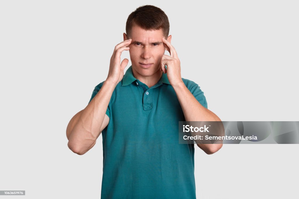 Stressful young man with dark hair, keeps index fingers on temples, suffers from headache, needs painkiller, dressed in casual t shirt, isolated over white background. Negative feeling concept Adult Stock Photo