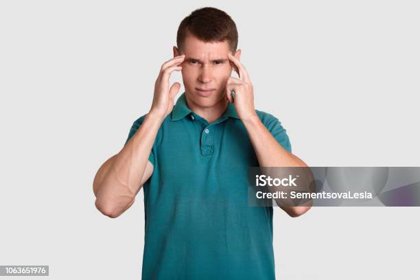 Stressful Young Man With Dark Hair Keeps Index Fingers On Temples Suffers From Headache Needs Painkiller Dressed In Casual T Shirt Isolated Over White Background Negative Feeling Concept Stock Photo - Download Image Now