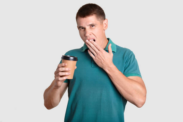 istock Tired guy yawns with sleepy expression, covers mouth with palm, holds disposable paper cup of coffee, dressed in casual t shirt, poses against white background. Morning time. Coffee break concept 1063652010