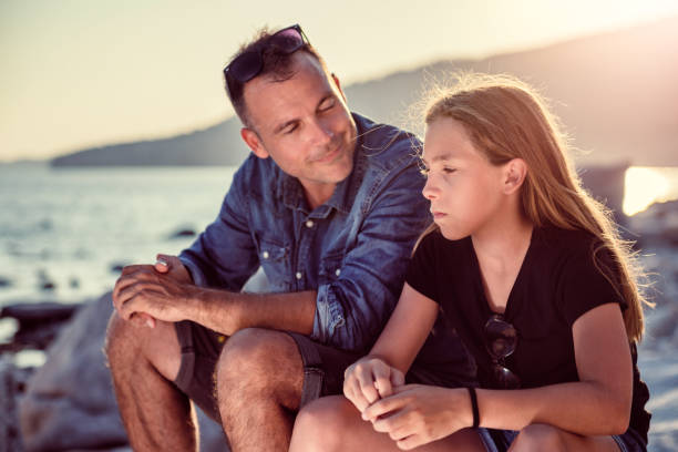 istock Father and daughter sitting on a rocky beach and talking 1063657548