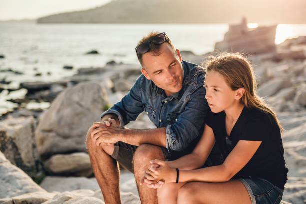 Father and daughter sitting on a rocky beach and talking Father and daughter relaxing on a rocky beach by the sea and having time together father and daughter stock pictures, royalty-free photos & images