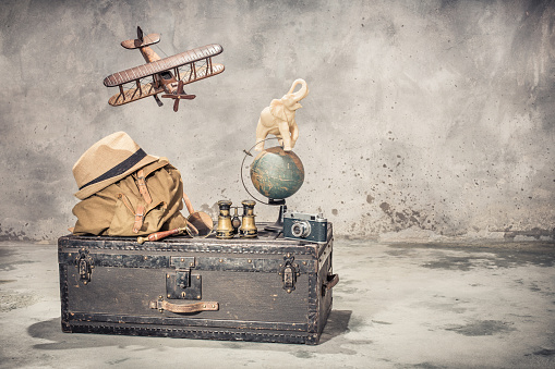 Vintage old classic travel trunk luggage, flying wooden toy plane, elephant on aged globe, tourist backpack, hat, binoculars, film camera, magnifier. Travel by air concept. Retro style filtered photo