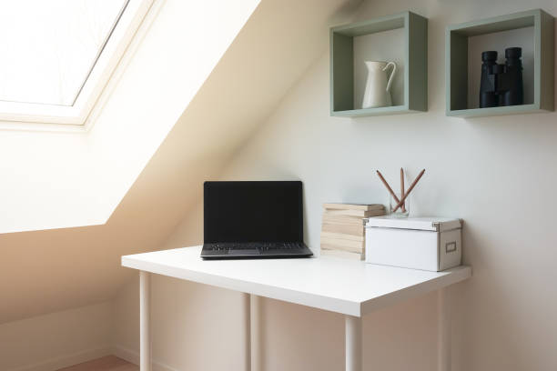 istock Simple workspace interior under skylight window in attic / loft. Laptop computer and a pile of books on a white modern office desk. 1063682542