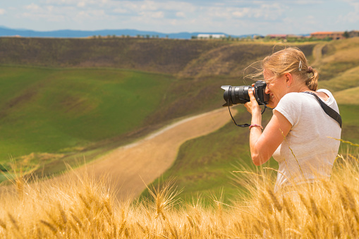 Senior woman photographing Toscana landscape, in wheat field with copy space in background. It is Italy, Europe.