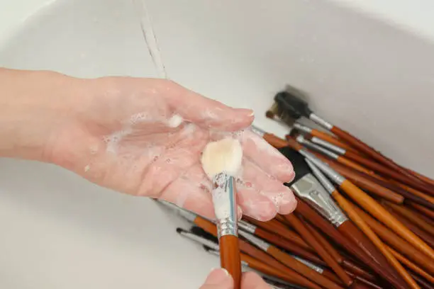 Photo of woman is washing dirty makeup brush with soap and foam in the sink