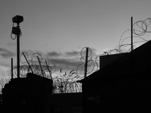Security camera mounted on a wall covered in razor wire in silhouette in monochrome against and evening shy