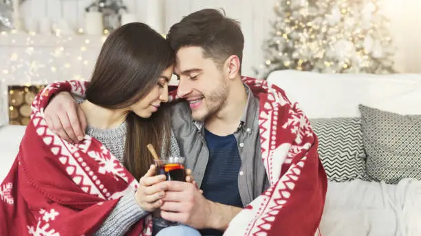 Loving couple embracing, covered with plaid and drinking mulled-wine against christmas tree