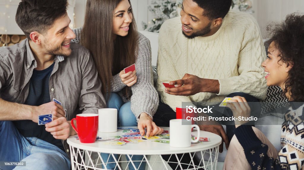 Happy friends playing game of cards UNO Multiracial friends having fun and playing game of cards UNO against christmas tree Playing Card Stock Photo