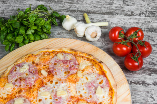 istock Thin pizza with ham, cheese and pineapple, rosemary and spices on a light wooden background. Italian pizza on a background of green basil and fresh vegetables 1063724100