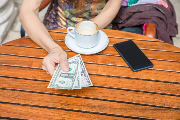 detail of woman hand with dollar bills paying a cappuccino coffee cup on brown wooden table, with black mobile phone