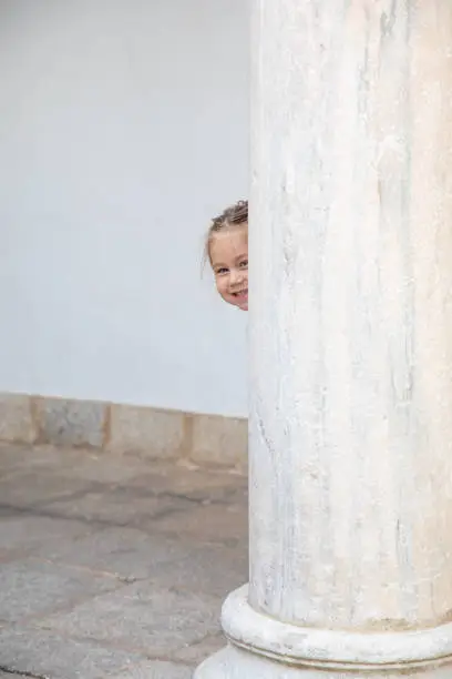 Photo of funny litlle child peeking and smiling behind ancient column