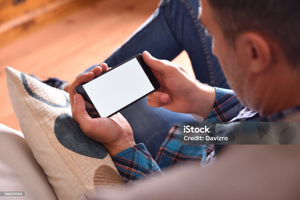 Man sitting on sofa watching multimedia content on a smartphone Adult man with blue shirt and jeans sitting on a sofa watching multimedia content on a smartphone. Raised viewpoint Horizontal Stock Photo