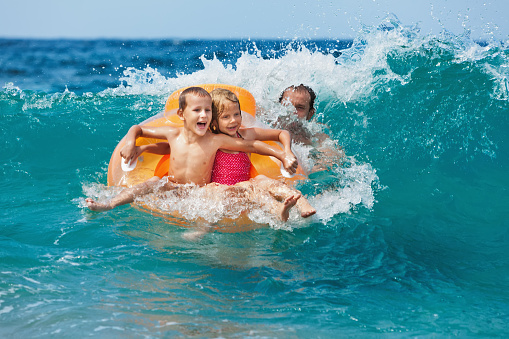 Happy kids have fun in sea surf on beach. Joyful couple of children on inflatable ring ride on breaking wave. Travel lifestyle, swimming activities in family summer camp. Vacations on tropical island