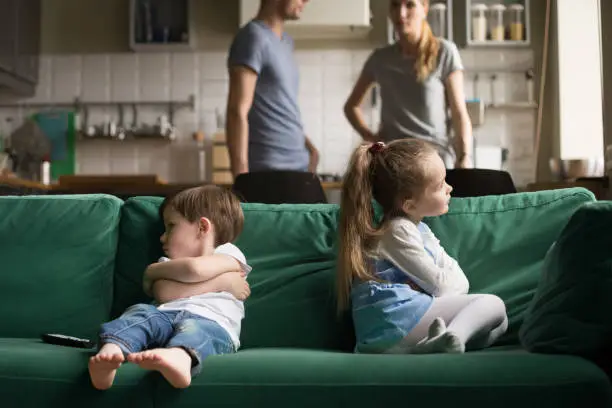 Upset offended toddler brother and sister sitting separately on couch, sofa with arms crossed, little girl and boy ignoring each other, not talking, puzzled parents discuss situation