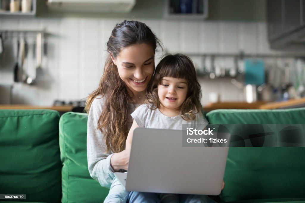 Happy laughing mother and daughter using laptop Happy laughing single mother and little preschool cute daughter using laptop online application, making video call, having fun, babysitter teaching small girl use computer, watching cartoons together Mother Stock Photo