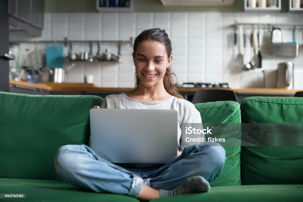 Happy smiling woman sitting on sofa and using laptop Happy smiling woman sitting on sofa, couch and using laptop at living room at home, watching funny video, learning language, video calling, mother working online Laptop Stock Photo