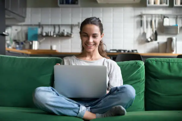 Happy smiling woman sitting on sofa, couch and using laptop at living room at home, watching funny video, learning language, video calling, mother working online