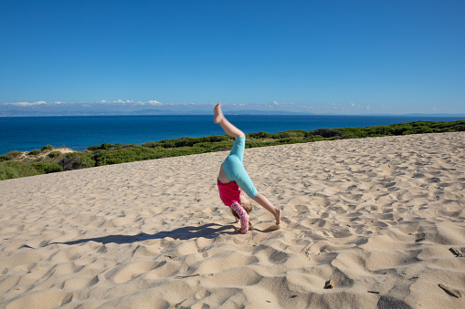 four years old girl doing the somersault or handstand on sand dune of Valdevaqueros (Tarifa, Cadiz, Andalusia, Spain), with ocean and Africa on the horizon