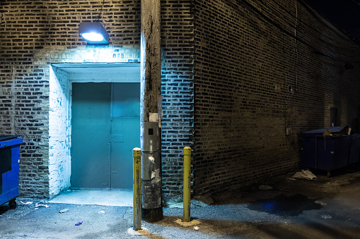 Dark and scary downtown urban city street corner alley with an eerie vintage industrial warehouse factory entrance and dirty dumpsters at night