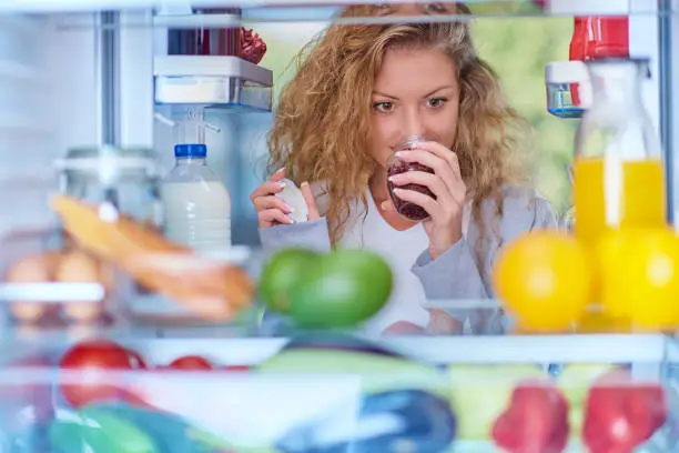 Woman smelling jam in front of fridge full of groceries. Picture taken from the inside of fridge.
