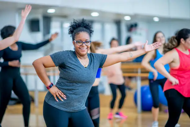 A woman of african descent looks into the camera smiling while she dances during a fitness class. She has one hand on her hip and the other arm extended out fully.