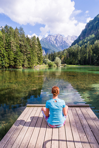 Young woman is sitting on a pier over the lake looking at the magnificent view  in Jezersko, Slovenia. Woman relaxing near the lake looking at the view. Travel adventure and freedom concept.