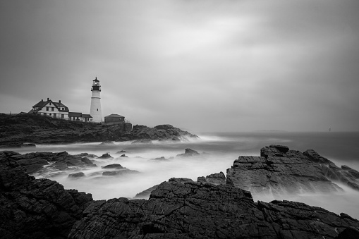 Long exposure black and white photo of the iconic Portland Head Light taken along the rocky coastline during a storm.