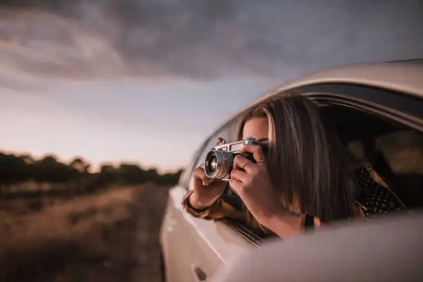 Photo of Young woman taking a picture with a vintage camera from car window