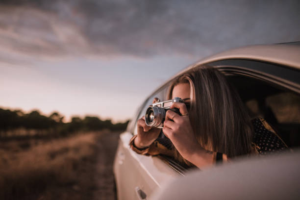 молодая женщина, снимавшая с винтажной камерой из окна автомобиля - photographer women retro revival camera стоковые фото и изображения