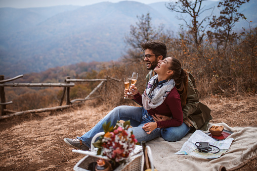 Couple at picnic. Couple sitting on blanket, laughing and drinking wine. Next to them picnic basket. Autumn time.