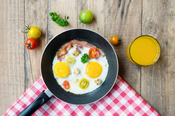 Fried eggs in a frying pan with cherry tomatoes and a glass of orange juice on the wooden table