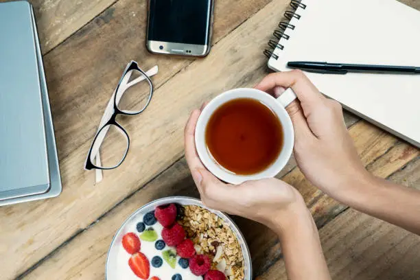 Business breakfast. Woman hands holding a cup of tea with yogurt and laptop computer on desk