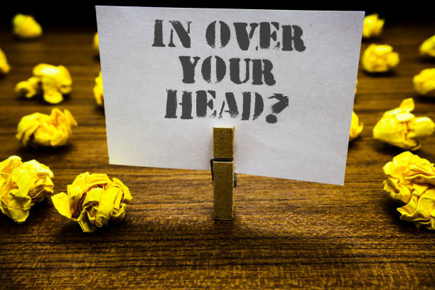 istock Text sign showing In Over Your Head question. Conceptual photo To be involved in a difficult situation problem Paperclip grip white page with grey text woody floor laid blurry yellow lob. 1063948666