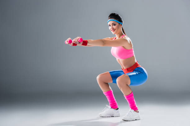sporty girl doing squats with dumbbells and smiling at camera on grey sporty girl doing squats with dumbbells and smiling at camera on grey 80s aerobics stock pictures, royalty-free photos & images