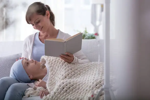 Smiling mother reading book to sick child with cancer wearing headscarf
