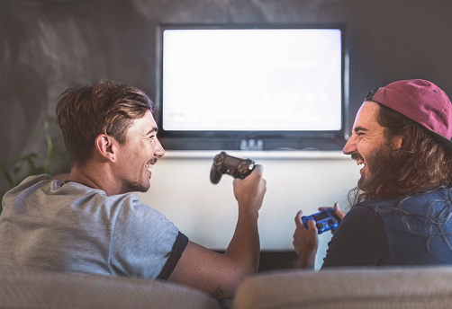 Rear view of cheerful young men playing video game on television at home. Male friends are laughing while sitting with game controllers on sofa. They are enjoying in living room.