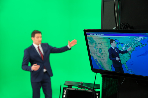Young weather man in front of a green background with small remote in his other hand. Weather forecast with map on the tv screen.