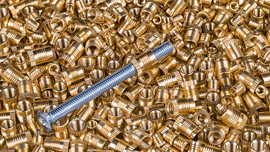 Close-up of a single long silvery-blue screw with countersunk-head. Bronze thread bushings. Pile of glossy metal parts. Gleaming fasteners. Idea of build, supply, store or assembly