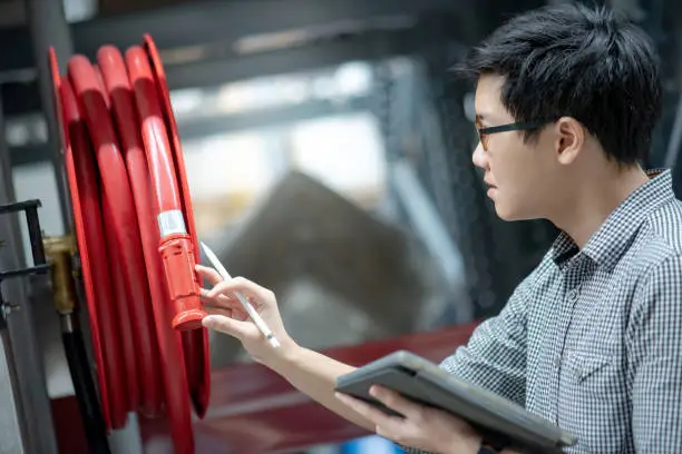 Young Asian male technician checking red fire hose reel by using digital tablet and pen. Building service and maintenance concepts