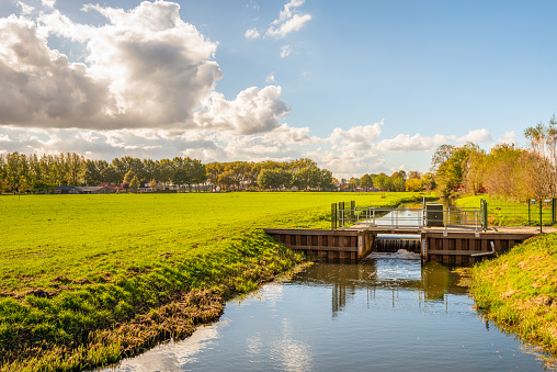 Dutch rural landscape in changing autumnal colors. In the foreground is a flowing weir in the stream with a smooth water surface. It is a sunny day with a blue sky and some clouds in the fall season.