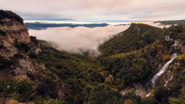 panoramic at dawn in a foggy dale. Waterfall falls through the precipice, forest has the first autumnal colors. In the background can see the mountains and the first light of sunrise. sky is cloudy