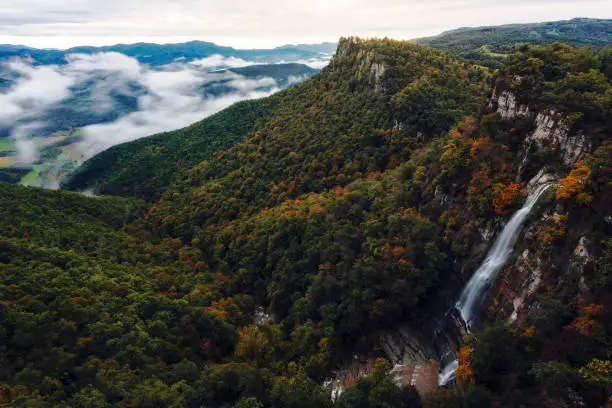 River falls through the stone wall of the mountain towards the valley during sunrise. The fog covers part of the fields. The leaves of some forest trees have the first red and yellow colors of autumn