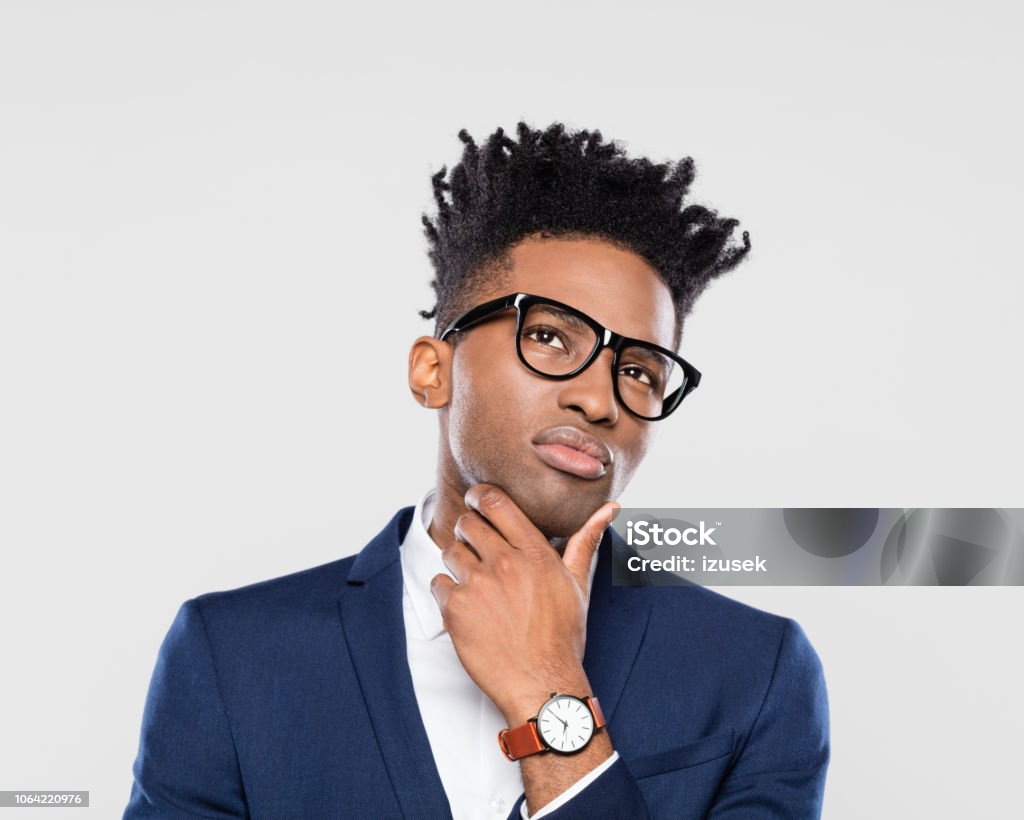Suspicious african businessman looks up doubtfully Suspicious young business man looks up doubtfully against gray background. Afro american man with stylish hairstyle, wearing eyeglasses looking upwards. Adult Stock Photo