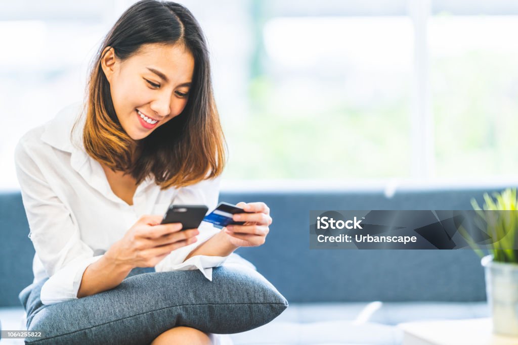 Young beautiful Asian woman using smartphone and credit card for online shopping at home with copy space. E-payment technology, shopaholic lifestyle, or mobile phone financial application concept Credit Card Stock Photo