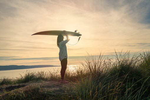 Woman holding a surfboard above her head at sunset, Sand dunes at Gwithian Towan beach, Cornwall.