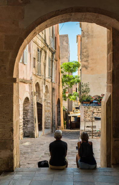 Two mature middle aged tourists take shelter from the hot midday sun Bonifacio, Corsica – September 30 2018: Two mature middle aged tourists take shelter from the hot midday sun under an arch on the steps of the Eglise Sainte Marie Majeure, looking down a narrow street of Bonifacio. corsica photos stock pictures, royalty-free photos & images