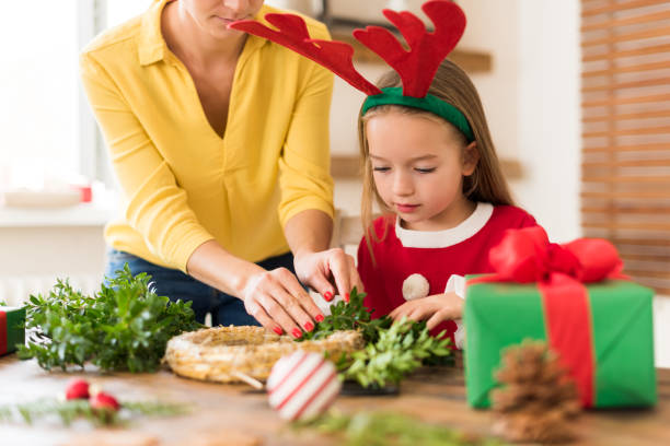 Cute preschooler girl wearing reindeer antlers and her mother making christmas wreath in living room. Christmas decoration family fun concept. stock photo