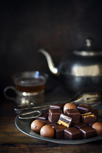 christmas chocolate sweets on a plate, silver teapot and cup blurry in the dark rustic background with copy space, vertical, selected focus, narrow depth of field