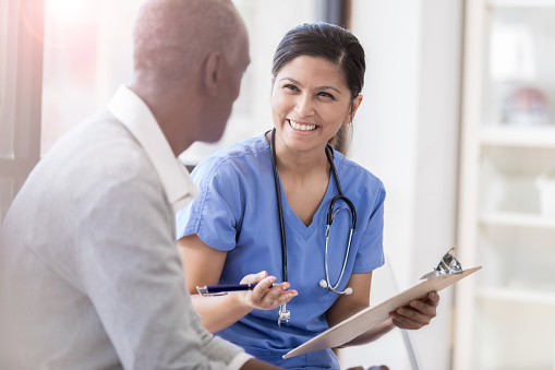 A smiling female doctor wears scrubs as she sits beside her unrecognizable senior male patient.  She gestures as she holds a clipboard and shares his test results.