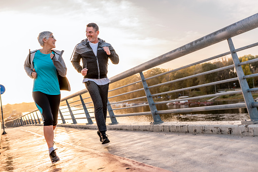 Photo of healthy Caucasian mature couple jogging in the city at early morning with a bridge clear sky as background. Man and gray hair woman exercising and jogging together in the city. Happy and smiling as they run along the path during sunset on a warm autumn day. Friendly competition.